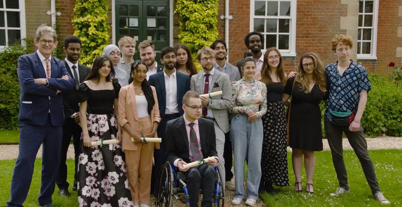 Group of diverse young adults and one older man posing outside a brick building covered in ivy, several holding rolled diplomas; a young man in a wheelchair sits at the front.