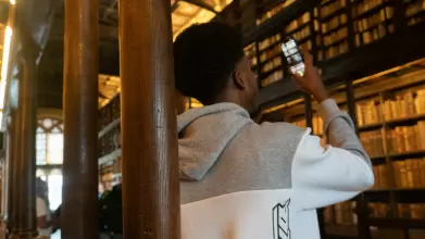 Person in a gray and white hoodie standing in a grand library with tall wooden columns and bookshelves. They are holding up a smartphone, possibly taking a photo or video. The library features an ornate ceiling, large windows letting in natural light, and
