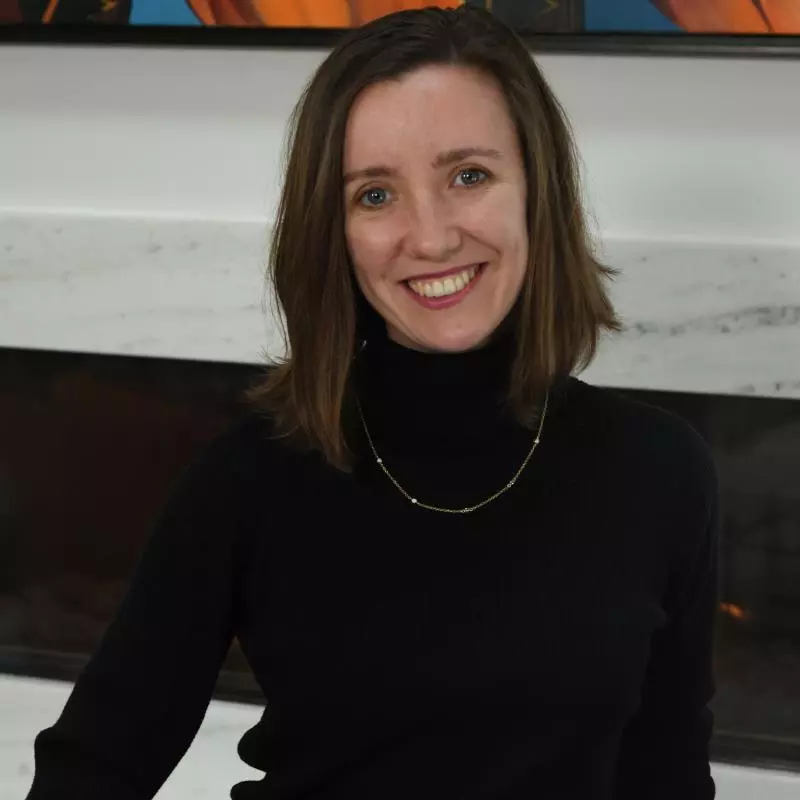 A young woman with shoulder length brown hair stands in a well-furnished room and smiles broadly