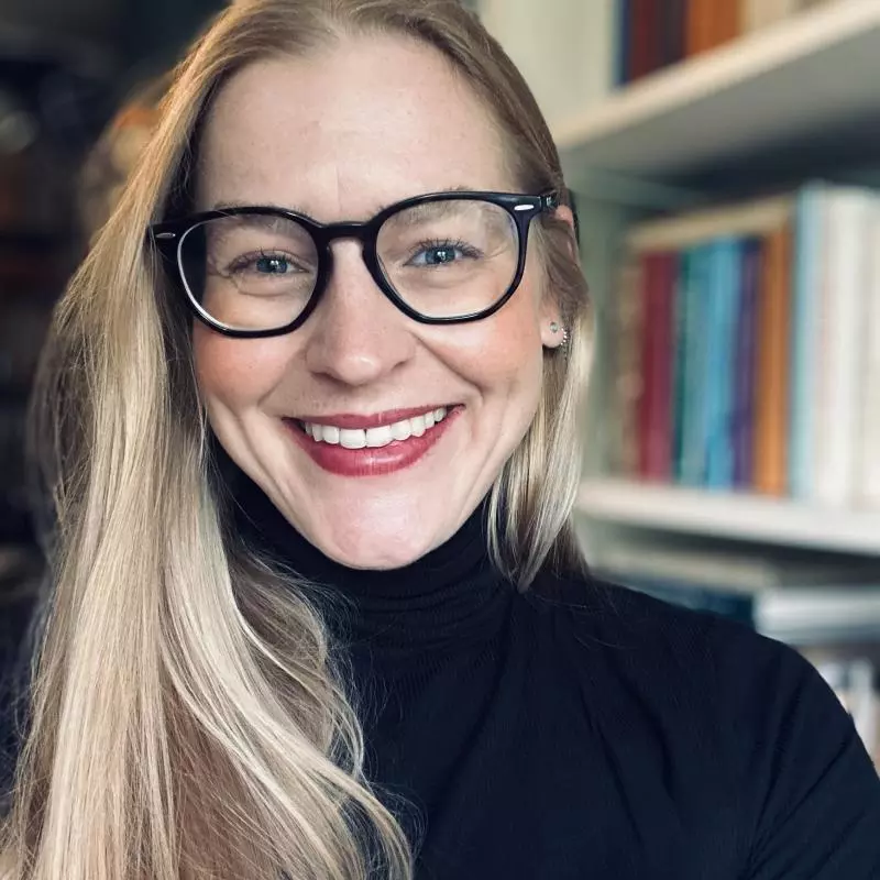 A young woman with long blonde hair and dark-rimmed glasses, smiling broadly