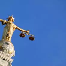 A picture of Lady Justice, a gold figure with arms outstretched holding a sword in one hand and scales in the other, against a clear blue sky